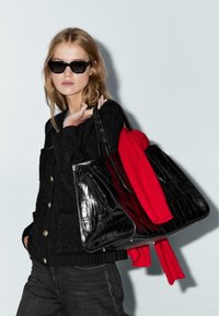 Donna che indossa occhiali da sole neri e un cardigan nero, mentre tiene una grande borsa nera con una trama, con un maglione rosso appoggiato sopra.