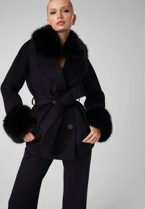 Femme portant un manteau noir ceinturé avec un large col en fourrure et des poignets en fourrure, associé à un pantalon noir, debout devant un fond neutre.