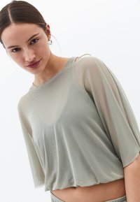 Lätt, genomskinlig topp i olivgrön med lösa ärmar och cropped fåll. Har en slät yta och subtilt vågmönster.