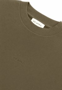 Κοντινή λήψη ενός ελαφρώς πρασινοκίτρινου μπλουζάκι με στρογγυλή λαιμόκοψη, που φέρει διακριτικό ανάγλυφο κείμενο και υφασμάτινη ετικέτα με "OH APRIL" στη λαιμόκοψη.