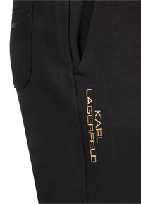Schwarze Sweatpants mit strukturierter Oberfläche, die über eine seitliche Tasche und eine goldene, gestickte "KARL LAGERFELD"-Markierung am Bein verfügen.