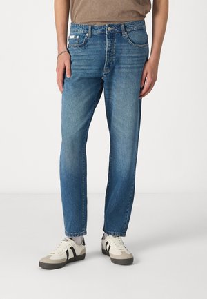 Jeans de denim azul con corte recto, acabado desgastado y diseño de cinco bolsillos. Combinados con zapatillas beige con rayas negras.