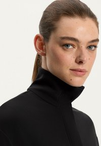 Zwarte hoge hals top met een gladde textuur, voorzien van een gedeeltelijke ritssluiting en een gestructureerde kraag. Model toont een neutrale uitdrukking.