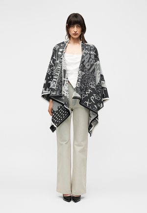 Žena nosí čierno-biely vzorovaný šál cez biely top, svetlobéžové široké nohavice a čierne lodičky so špicatou špičkou, stojaca na bielom pozadí.