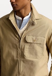 Muž v béžové zipové bundě přes krémový pletený svetr a košili s modro-bílými pruhy a zapínáním na knoflíky.