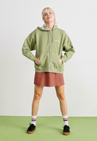 Zelená mikina na zips s predným vreckom a šnúrkami, kombinovaná s sukňou v farbe hrdze, bielymi ponožkami s červenými a čiernymi pruhmi a čiernymi topánkami.