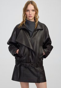 Giacca di pelle nera con vita con cintura, design over size, chiusura con zip e colletto alto. Texture liscia con tasche laterali.