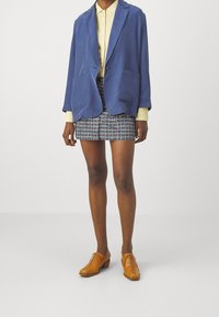 Modrý sametový blazer se dvěma předními kapsami, v kombinaci s kostkovanou mini sukní a hnědými koženými šněrovacími botami. Pod ním žlutá košile na knoflíky.