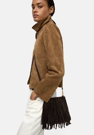 Kobieta nosząca brązową zamszową kurtkę i białe spodnie, niosąca ciemnobrązową torbę na ramię z frędzlami, patrząca na bok na jednolitym tle.