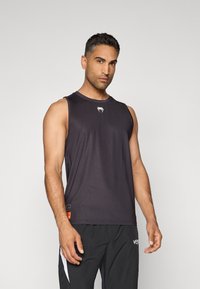 T-shirt de sport sans manches noir en tissu léger et lisse avec un col rond, présentant un logo blanc sur la poitrine et une étiquette orange.
