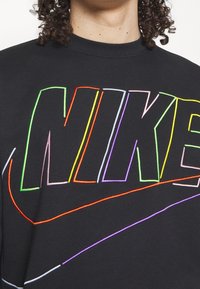 Personne aux cheveux bouclés portant un sweat-shirt noir avec un grand logo NIKE multicolore en contour sur la poitrine.