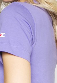 Lila bomulls t-shirt med korta ärmar och rundad halsringning. Har en liten röd och vit logotyp på vänster ärm. Slät textur.