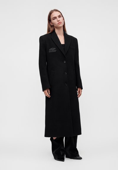 Jeune femme aux longs cheveux raides portant un long manteau noir à boutons et un large pantalon noir sur un fond clair uni.