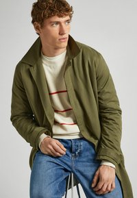 Olijfgroene lange jas over een wit truitje met rode strepen, gecombineerd met blauwe spijkerbroek. Heeft een brede kraag en geribbelde manchetten.