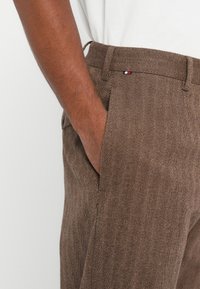 La mano di un uomo nella tasca di pantaloni marroni a motivo herringbone, indossati con una camicia bianca, che mostrano i passanti per la cintura e una piccola etichetta a bandiera.