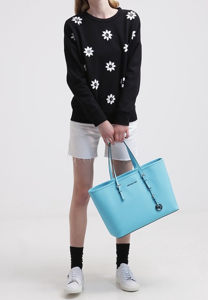 Personne portant un pull noir avec des fleurs blanches, un short blanc, des chaussettes noires, des baskets blanches, tenant un grand sac à main bleu clair.