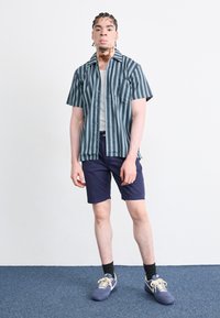 Chemise à manches courtes à boutons à rayures teal et marine, associée à un short marine. La chemise possède une poche poitrine. Portée avec des baskets marines.
