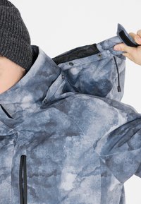 Sininen-harmaa kuvioitu takki korkealla kauluksella, neppareilla ja säädettävällä hupulla. Ominaisuuksiin kuuluu sileä pinta ja vetoketjulliset sivutaskut.