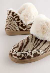 Chaussons doublés en fausse fourrure avec un motif animalier beige et marron, présentant des coutures blanches et une semelle en caoutchouc flexible.