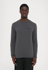 Young man wearing a fitted dark gray long-sleeve shirt with "EA7 Emporio Armani" logo on the left chest against a plain background.