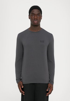 Young man wearing a fitted dark gray long-sleeve shirt with "EA7 Emporio Armani" logo on the left chest against a plain background.