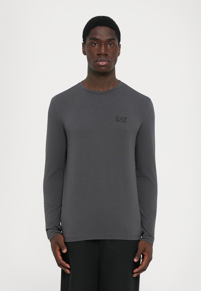 Young man wearing a fitted dark gray long-sleeve shirt with "EA7 Emporio Armani" logo on the left chest against a plain background.