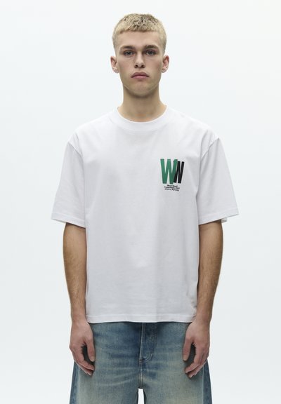 White cotton T-shirt with short sleeves, featuring a green and black graphic 'WW' logo on the left chest. Worn with faded denim shorts.