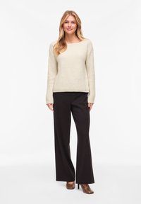Πουλόβερ σε ανοιχτό μπεζ χρώμα, πλεκτό, με μακριά μανίκια και λαιμόκοψη σκάφος, συνδυασμένο με ψηλόμεσα μαύρα παντελόνια φαρδιάς γραμμής και καφέ τακούνια.