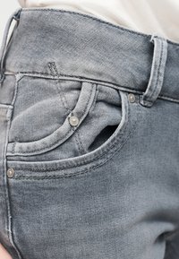 Gros plan sur une personne portant un jean en denim gris clair avec poche avant visible, passants de ceinture et détails de rivets argentés.