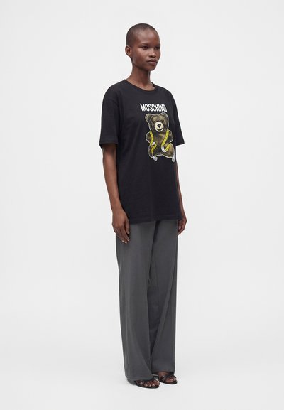 Άτομο που φοράει μαύρο t-shirt Moschino με γραφικό αρκουδάκι, γκρι παντελόνι με φαρδιά μπατζάκια και μαύρα ανοιχτά σανδάλια, στέκεται σε απλό φόντο.