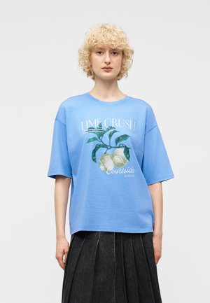 Persoană cu păr scurt, blond și creț, purtând un tricou albastru cu imprimeu "Lime Crush" și o fustă plisată închisă la culoare, stând pe un fundal simplu.