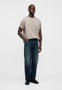 Άνδρας που φοράει μπεζ πουκάμισο με γυρισμένα μανίκια, σκούρο μπλε τζιν με ελαφριά φθορά και μαύρα αθλητικά παπούτσια, στέκεται με το ένα χέρι στην τσέπη.