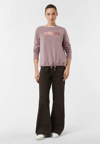 Jasnofioletowy sweatshirt z metalicznym różowym hasłem "LESS IS MORE" z przodu, połączony z ciemno-brązowymi spodniami dzwonami. Teksturowany materiał, talia ze sznurkiem.