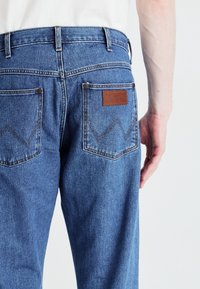Modri kavbojki iz denima z rjavim usnjenim oznako z blagovno znamko "Wrangler", značilnim šivanjem zadnjih žepov v vzorcu zigzag in klasičnim krojem.