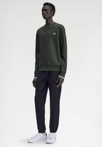 Felpa verde con logo bianco, abbinata a pantaloni neri a sigaretta e sneakers bianche. La felpa presenta polsini e orlo a costine.