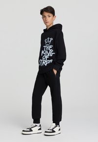 Felpa nera con testo bianco "IL RE DELLA STRADA," abbinata a pantaloni jogger neri e sneaker bianchi e neri. Tessuto morbido, vestibilità rilassata.