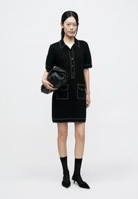 Robe noire à manches courtes, boutonnée à l'avant, avec des surpiqûres blanches contrastantes, deux poches décoratives, associée à un sac à main noir et des chaussettes noires.