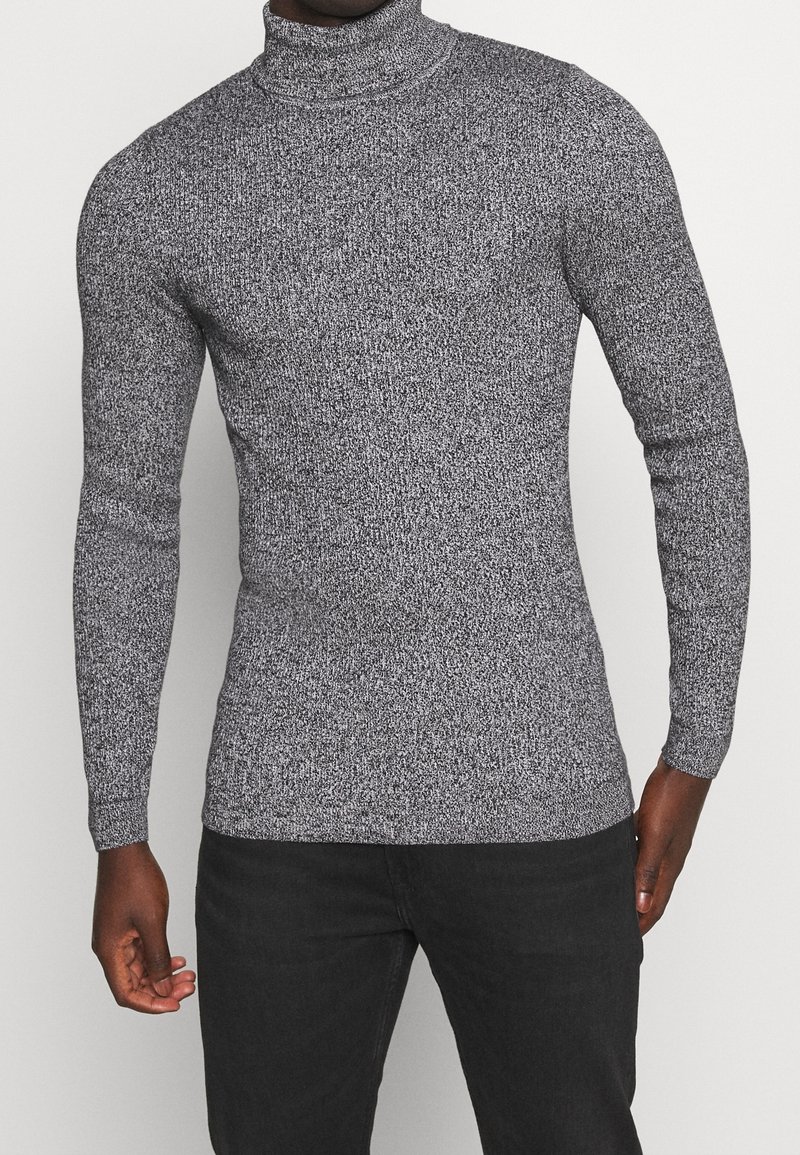 Homme portant un pull à col roulé ajusté gris et noir moucheté à manches longues et un pantalon noir, sur fond uni.