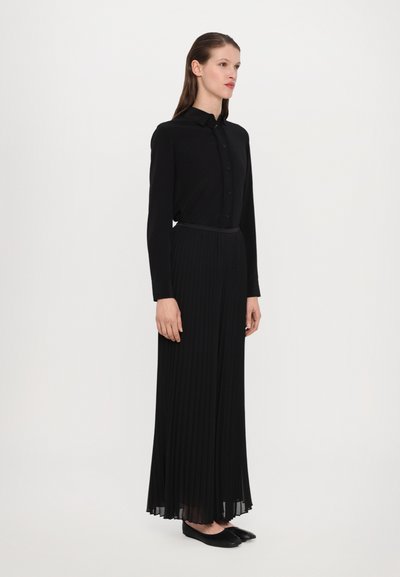Čierna košeľa s dlhými rukávmi a gombíkovým zapínaním spolu s čiernou maxi sukňou s plisovaním. Úplný outfit dopĺňajú ploché čierne topánky. Hladká textúra, minimalistický dizajn.