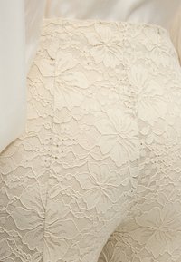 Κοντινό πλάνο μπεζ δαντέλας με λουλουδένια σχέδια σε ένδυμα, που δείχνει λεπτομερή υφή και ραφές.