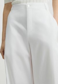 Weiße hochtaillierte Hose aus glattem Stoff, mit einer Vorderentasche und Knopfverschluss. Taillierte Passform mit klaren Falten.