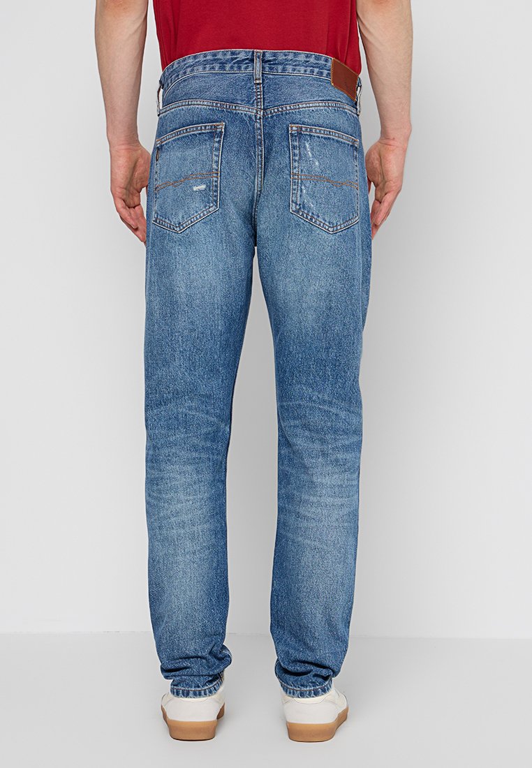 Mann in verblassten blauen Jeans und weißen Sneakers, der gegen eine schlichte graue Wand steht, Blick von hinten.