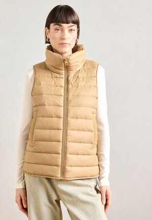 Gilet puffo beige con collo alto, design trapuntato, zip frontale e tasche laterali, abbinato a un top bianco a maniche lunghe e jeans chiari.