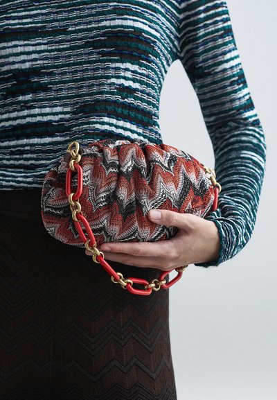 Person med en teal randig topp och en brun zigzag-mönstrad kjol håller en röd, svart och vit zigzag-mönstrad clutch med en röd och gyllene kedjesnodd.