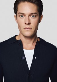 Granatowy kardigan z dekoltem w szpic, teksturowanym splotem i trzema guzikami. Noszony na białej koszulce z okrągłym dekoltem, ukazujący prosty, czysty design.