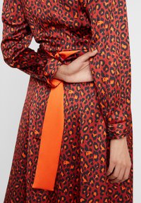 Personne portant une robe imprimée léopard rouge et orange avec une ceinture orange vif nouée à la taille, une main reposant sur le dos.