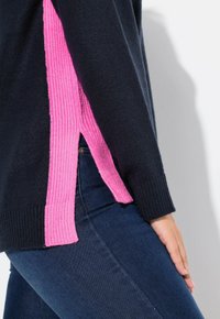 Pull en tricot avec un corps bleu marine et des panneaux latéraux côtelés rose fluo. Manches longues, fentes sur les côtés et finition texturée. Associé à un denim foncé.