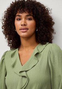 Blouse verte et texturée avec un décolleté en V, des accents volants et des manches bouffantes. Un motif tissé prononcé renforce l'intérêt visuel.