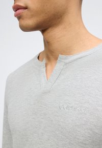 Κοντινό πλάνο ενός ατόμου που φοράει ανοιχτό γκρι μακρυμάνικη μπλούζα με υφή, ντεκολτέ με σκίσιμο και διακριτική κεντημένη λογότυπο Calvin Klein.