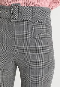 Grijze geruite broek met een gestructureerde stof, brede riem en riemlussen. Het patroon combineert kleine en grote ruiten met houndstooth accenten.
